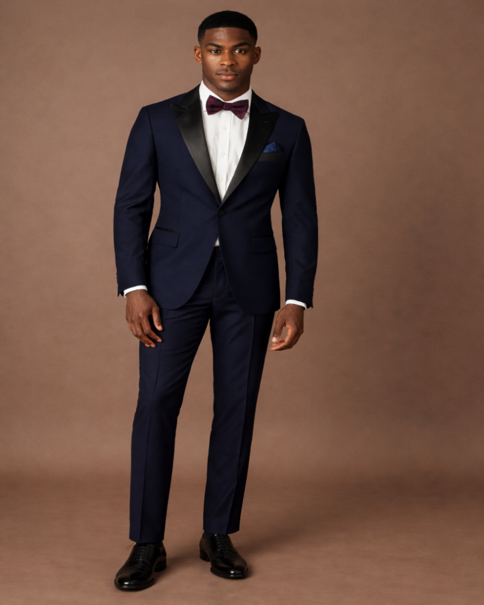 Man wearing a navy blue tuxedo with a white shirt and black shoes on a brown background